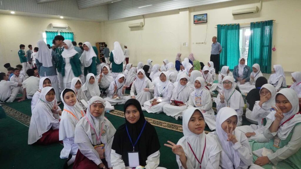Murid-murid perempuan baru yang mengenakan hijab tersenyum ceria sambil duduk lesehan mengikuti rangkaian acara pengenalan lingkungan sekolah.